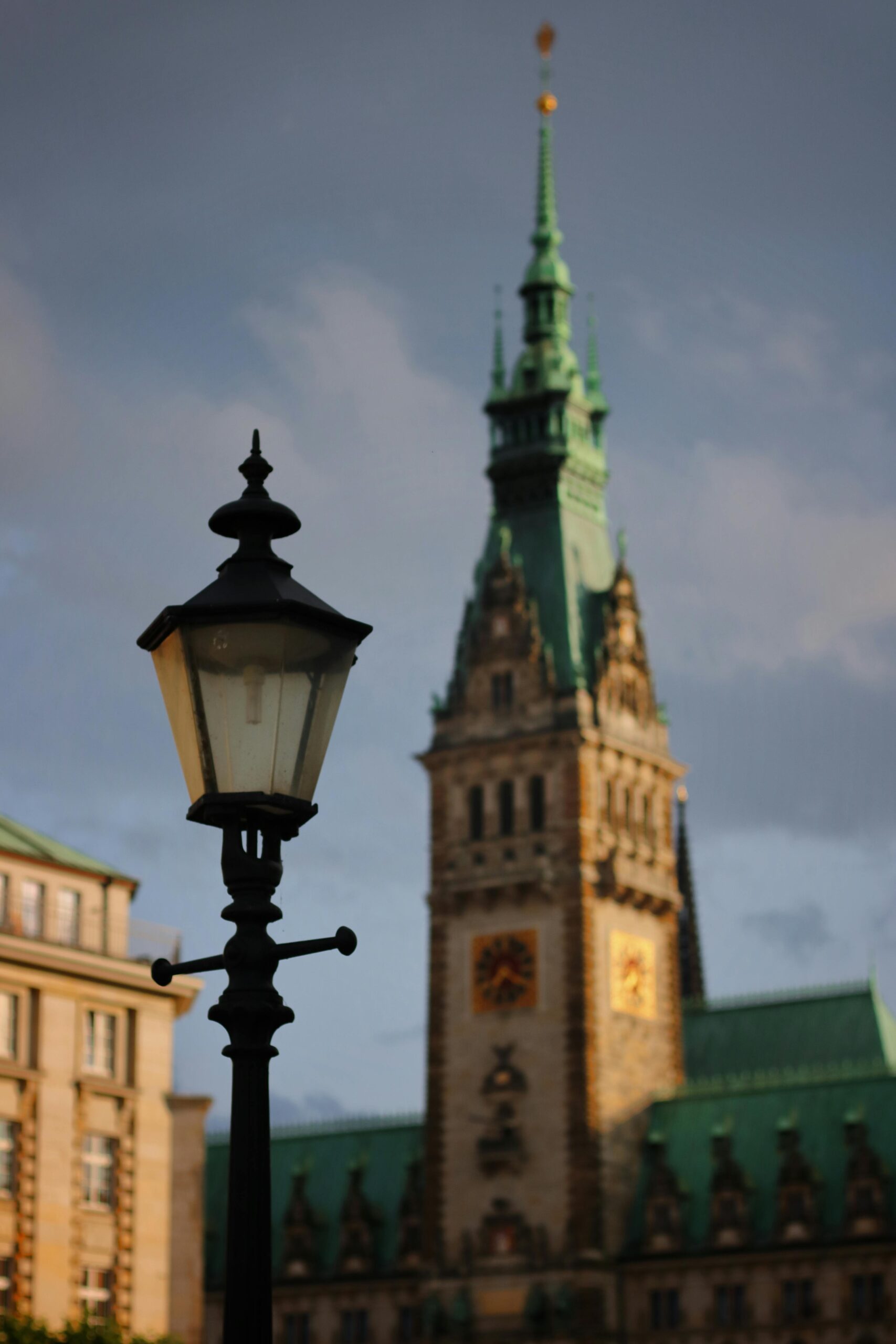 Historic lamppost with blurred background of Hamburg's Town Hall tower under a cloudy sky.